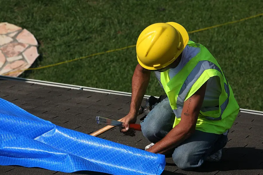 Worker fastening a protective tarp over a shingle roof after storm damage